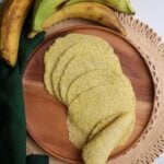 Plantain Rotis (tortillas) on a wooden plate with a few plantains above the plate and a green napkin to the left