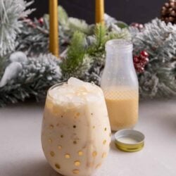 Ponche de creme in a glass with a bottle behind it and Christmas decor in the background.