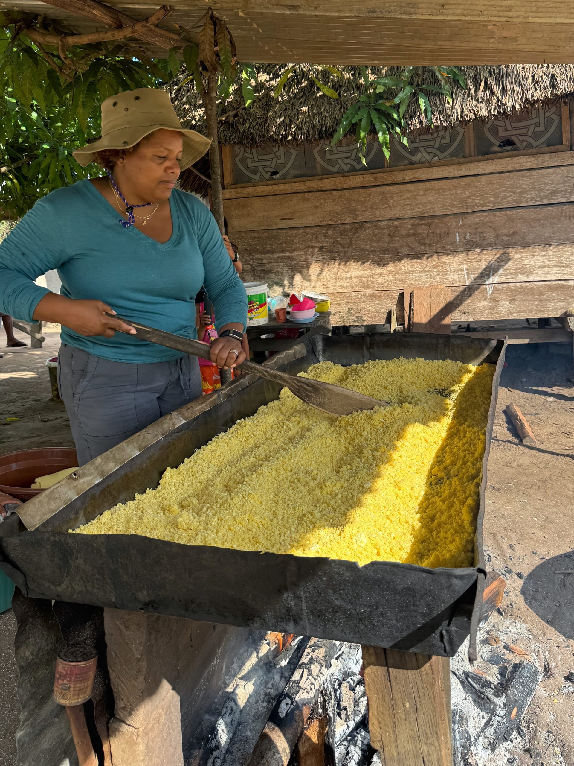 A woman wearing a sunhat and long-sleeve shirt toasts grated cassava over an open fire in a large rectangular metal pan, surrounded by a village setting with wooden structures and trees.