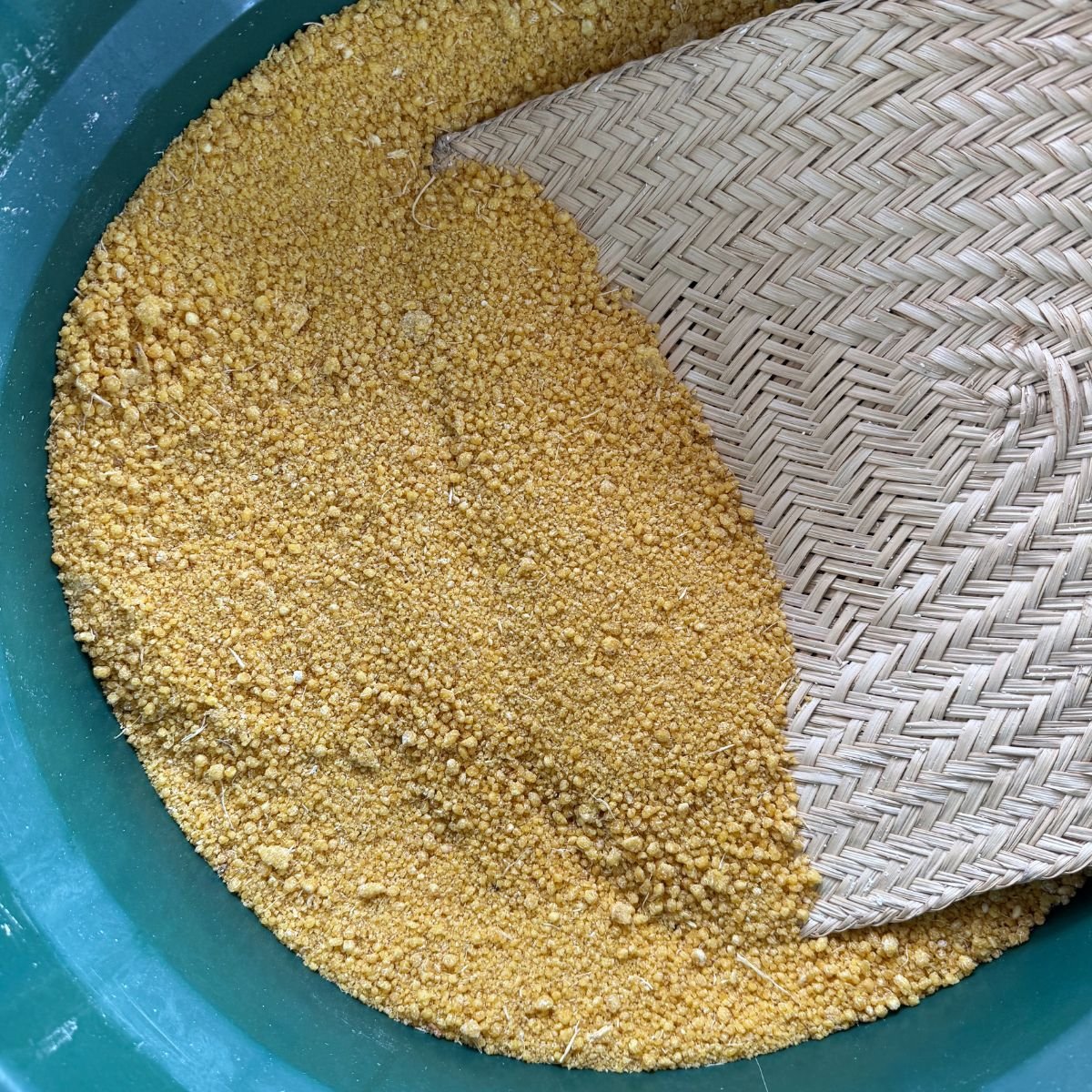Close-up of golden yellow farine in a green basin, partially covered by a woven straw sifter used during the cassava processing.