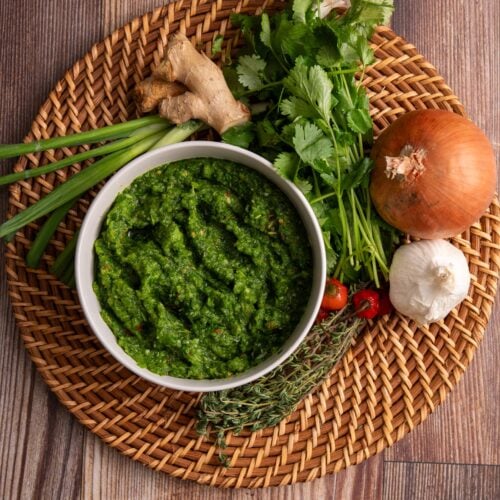Blended green seasoning in a bowl resting on a woven plate with aromatics