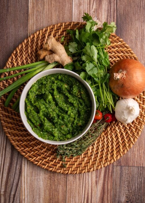 Blended green seasoning in a bowl resting on a woven plate with aromatics
