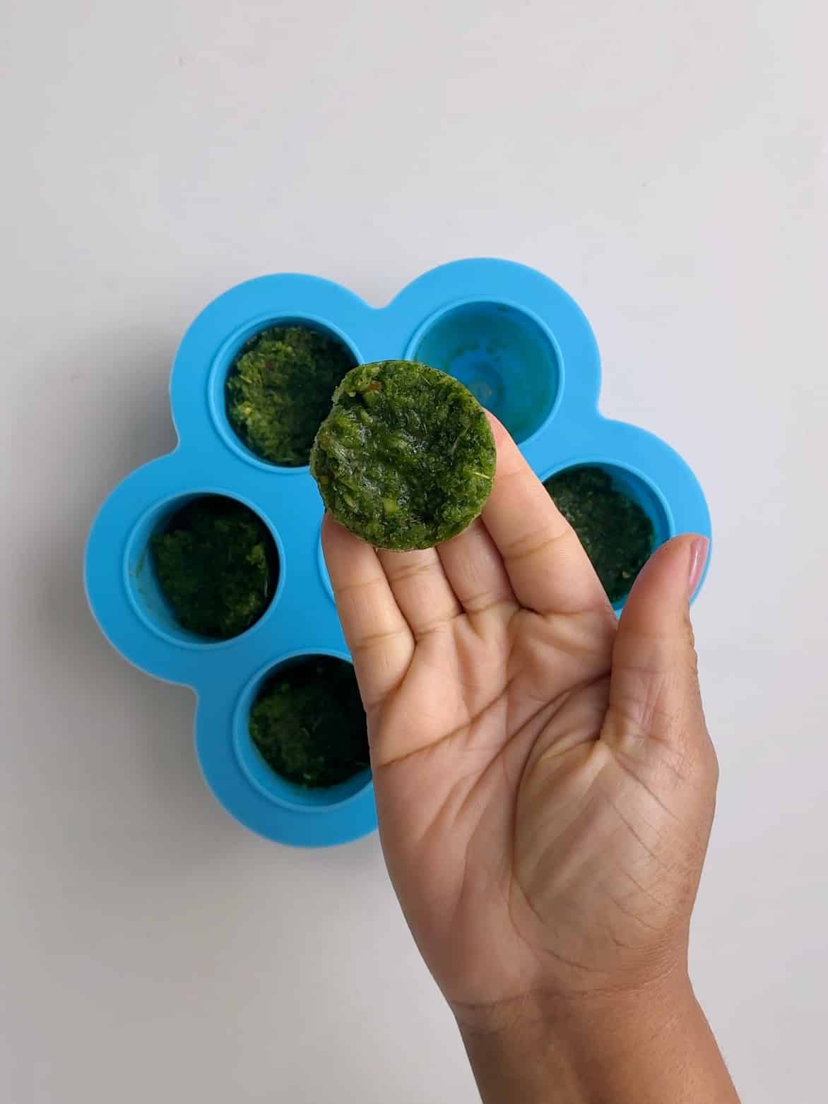 Frozen green seasoning in a silicone mold. One ball is out of the mold and resting on stretched out finger tips above the mold