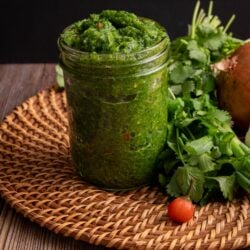 Green seasoning in a mason jar. The jar is on a woven plate and there are aromatics to the right of the jar.
