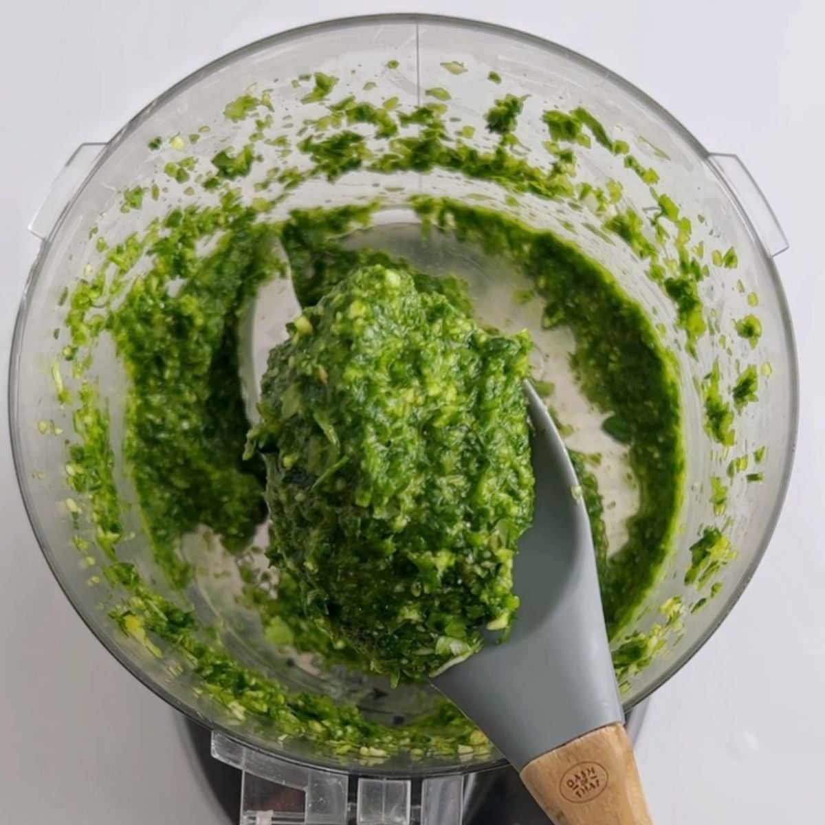 A thick, bright green herb paste being scooped from a food processor bowl with a spatula, showing the finished green seasoning texture.