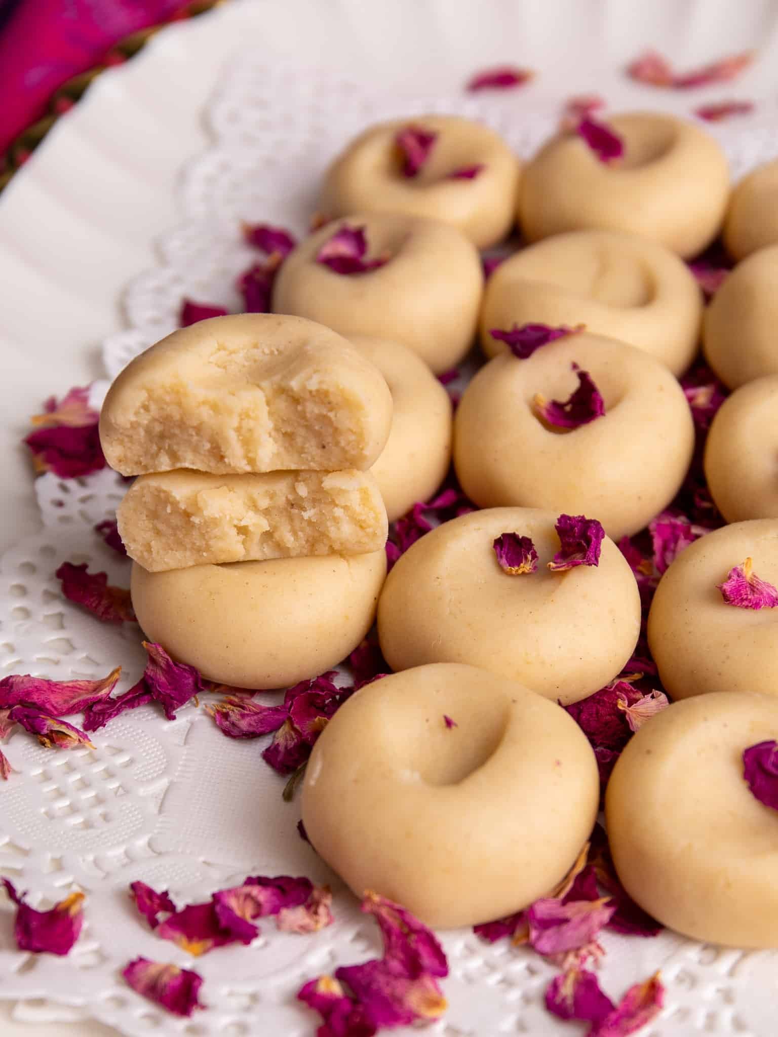 Close-up of Guyanese Milk Peera arranged on a white lace-lined platter and sprinkled with dried rose petals. One peera is cut in half and stacked on top, showing its soft, slightly crumbly interior.