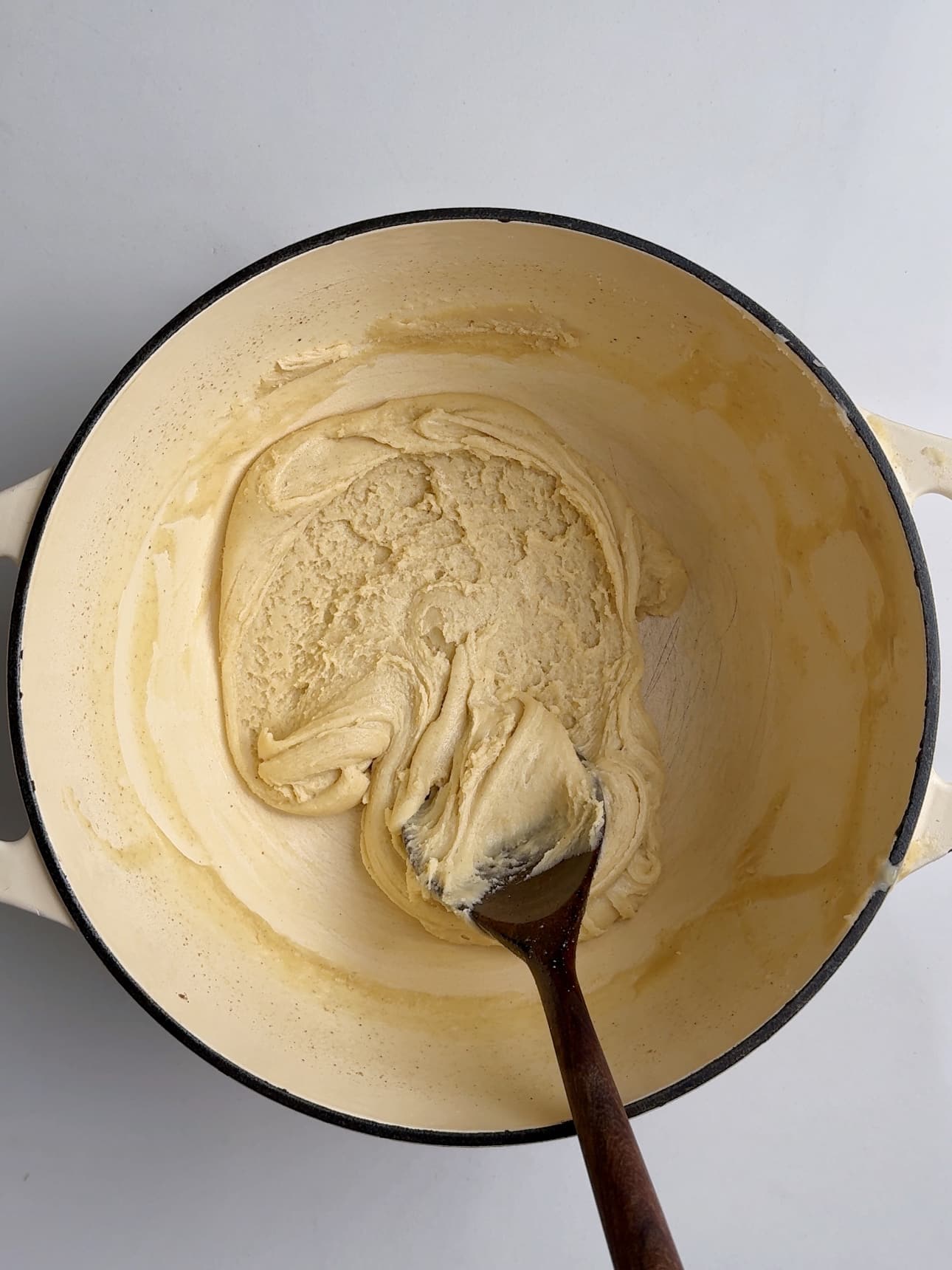 Overhead view of a cream-colored Dutch oven containing a thick, dough-like milk mixture that has fully reduced. A wooden spoon lifts and folds the dense paste, which holds its shape and pulls cleanly from the sides of the pot—showing the finished stage before shaping into Guyanese Milk Peera.