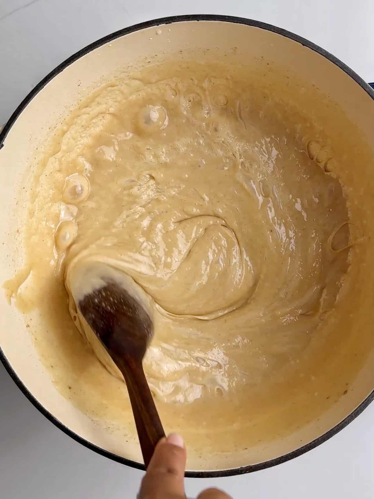 Overhead view of a cream-colored Dutch oven with a wooden spoon stirring a thick, smooth milk mixture that has reduced to a glossy caramel color. The mixture pulls away from the sides of the pot as it’s stirred, showing the final thickening stage of making Guyanese Milk Peera