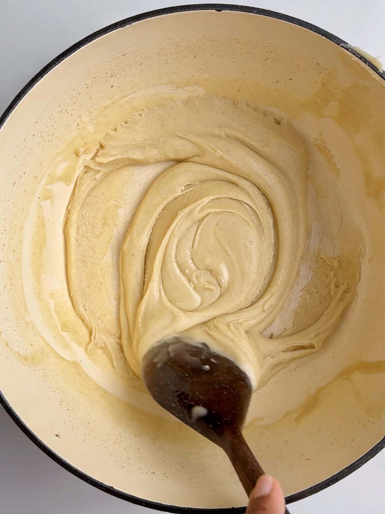 Overhead view of a cream-colored Dutch oven with a wooden spoon stirring a very thick, smooth milk mixture that has reduced to a pale caramel paste. The mixture forms soft folds and swirls and pulls cleanly from the sides of the pot, indicating it is ready to be shaped into Guyanese Milk Peera.