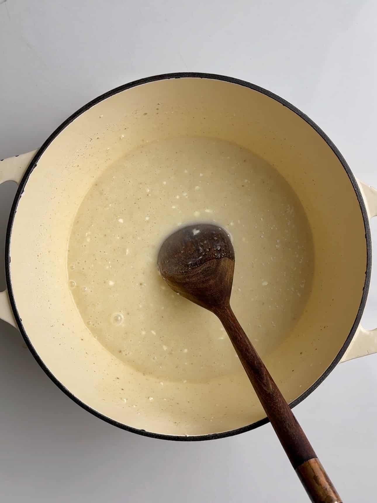 Overhead view of a cream-colored Dutch oven filled with a pale, milky mixture dotted with small curds. A wooden spoon rests inside the pot, partially submerged in the simmering mixture against a light background.