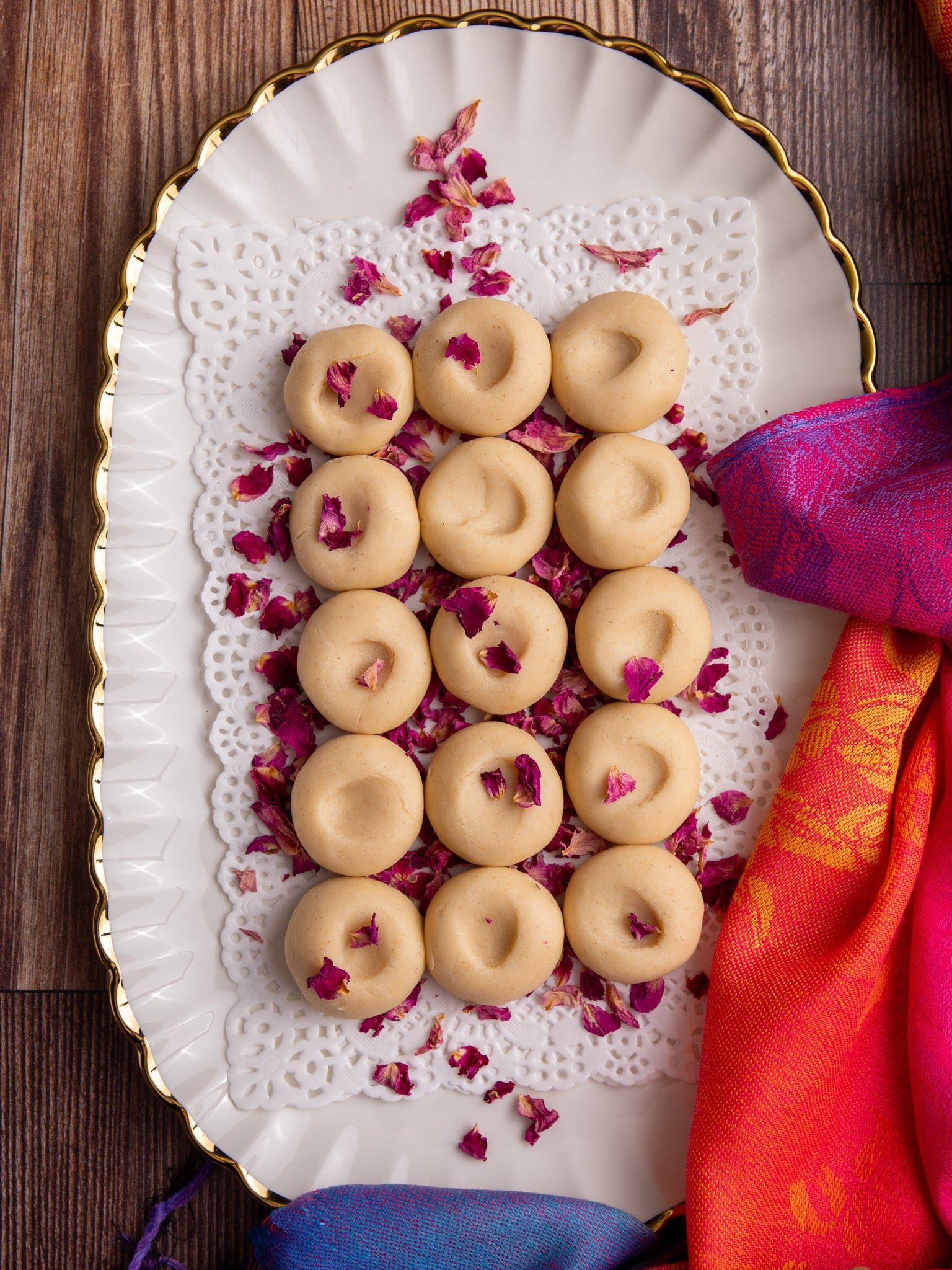 A white scalloped serving platter with gold trim holds neatly arranged round Guyanese pera sweets, each with a small thumbprint indentation in the center. The pera are placed on a white lace doily and sprinkled with dried pink rose petals. The platter sits on a wooden surface, partially framed by bright pink, orange, and blue fabric.