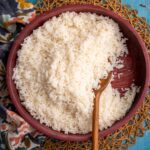 Serving bowl of coconut rice with a wooden spoon on a woven placemat over a blue wooden surface.