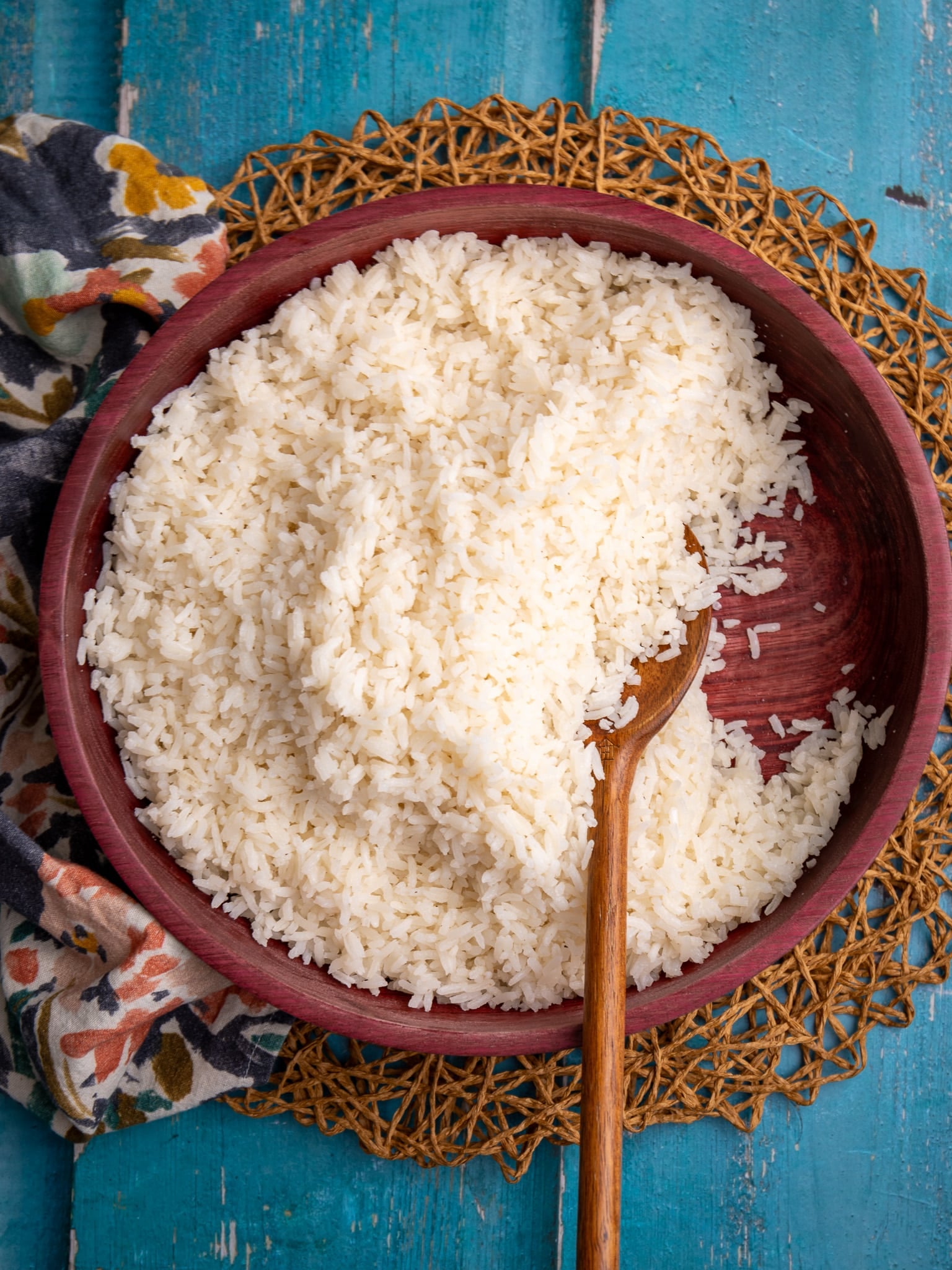 Serving bowl of coconut rice with a wooden spoon on a woven placemat over a blue wooden surface.