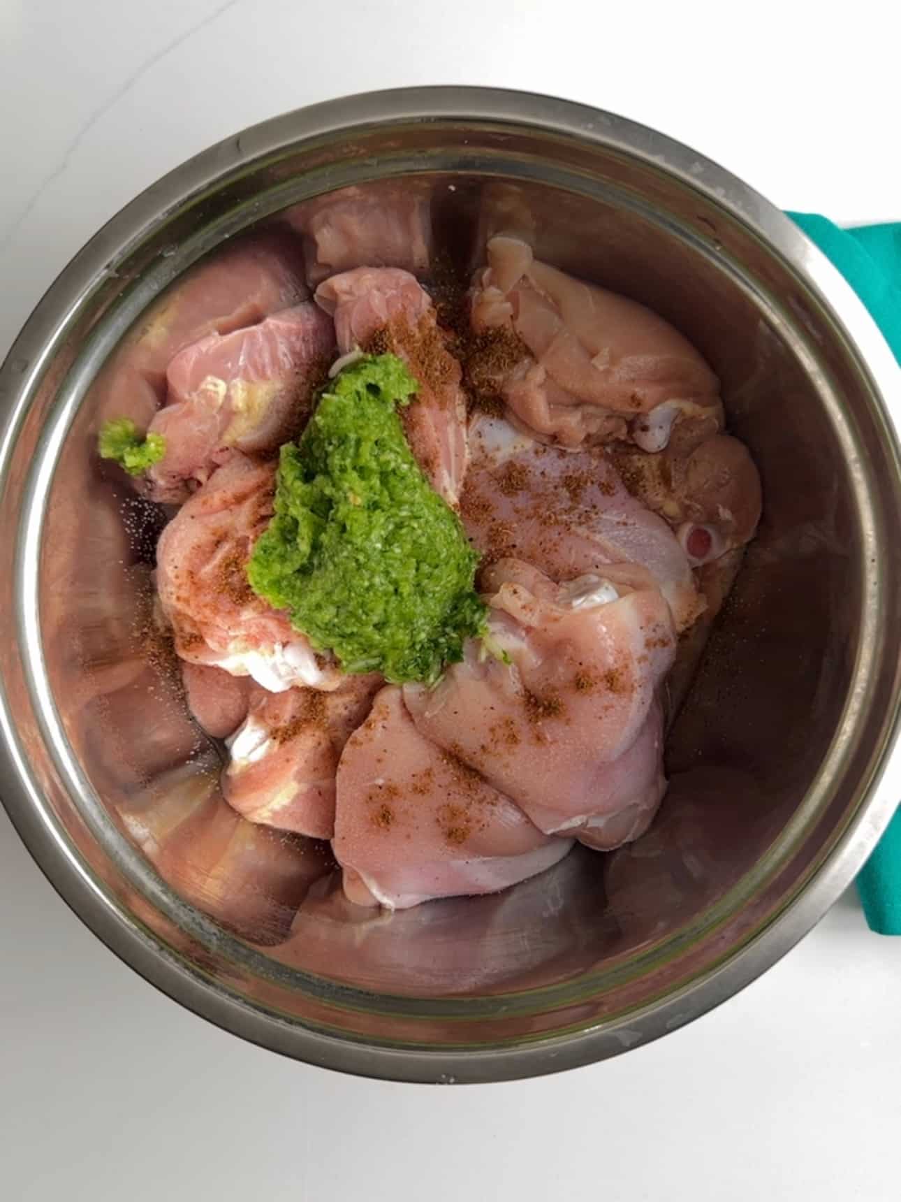 Raw chicken in a metal bowl seasoned with spices and a green herb seasoning paste.