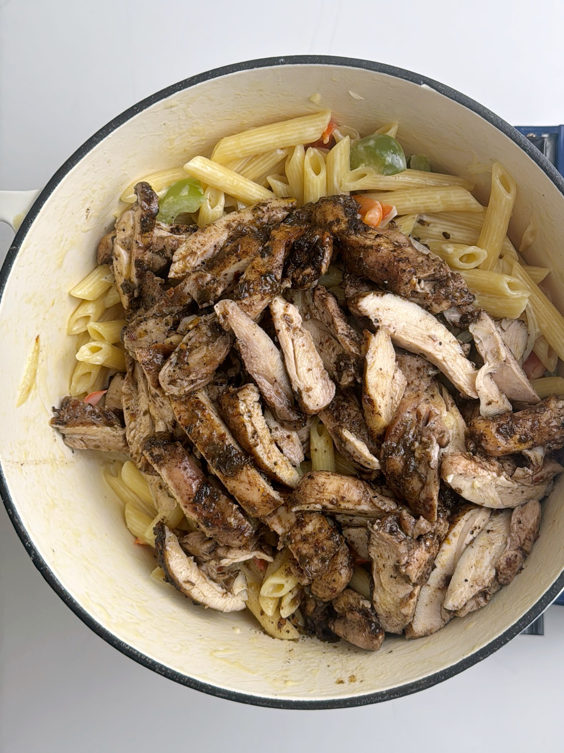Cooked sliced jerk chicken resting on top of creamy pasta with bell peppers in a large pot.