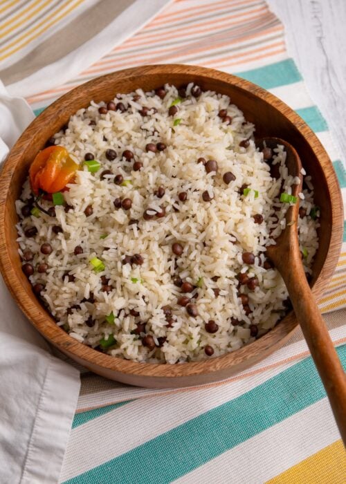 A wooden bowl filled with fluffy Pigeon peas and rice, garnished with chopped scallions and a whole Scotch bonnet pepper on top. The bowl sits on a striped cloth with a wooden spoon resting inside, highlighting the simple, comforting Caribbean rice dish