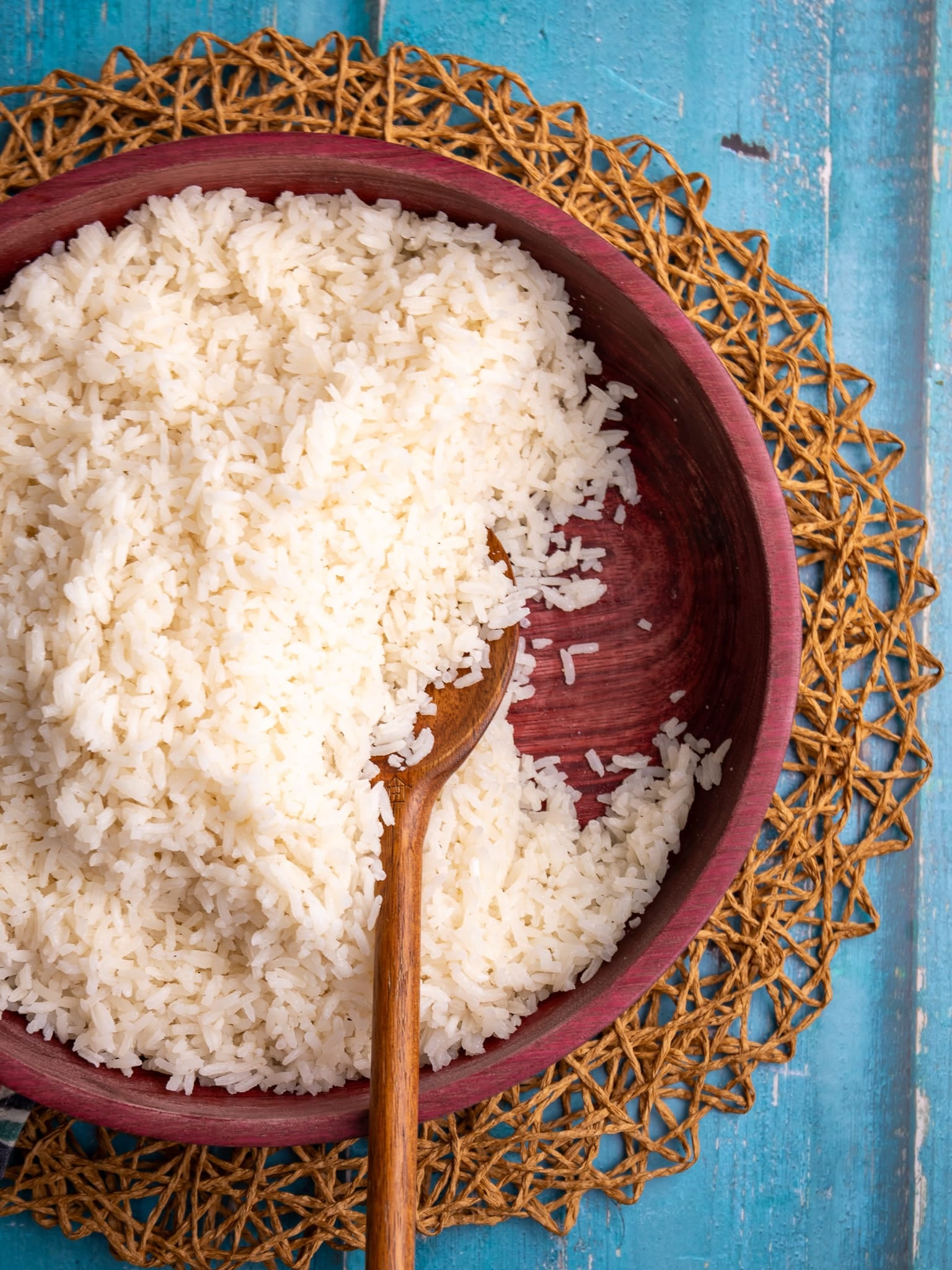 Serving bowl of coconut rice with a wooden spoon on a woven placemat over a blue wooden surface.
