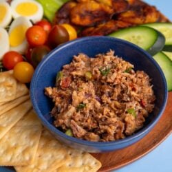 A bowl of sardines served with crackers, sliced cucumbers, cherry tomatoes, boiled eggs, and fried plantains on a plate.
