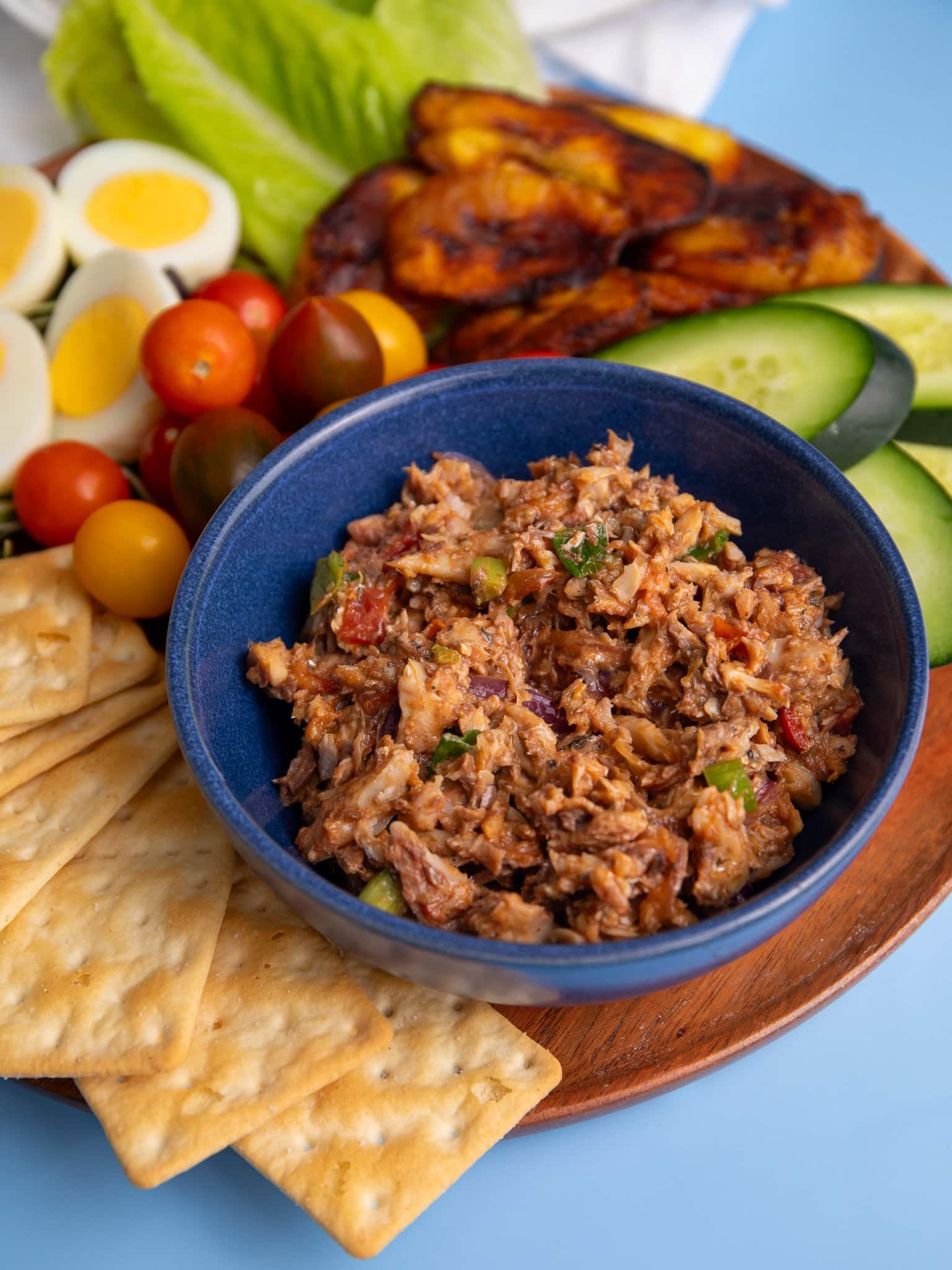 A bowl of sardines served with crackers, sliced cucumbers, cherry tomatoes, boiled eggs, and fried plantains on a plate.