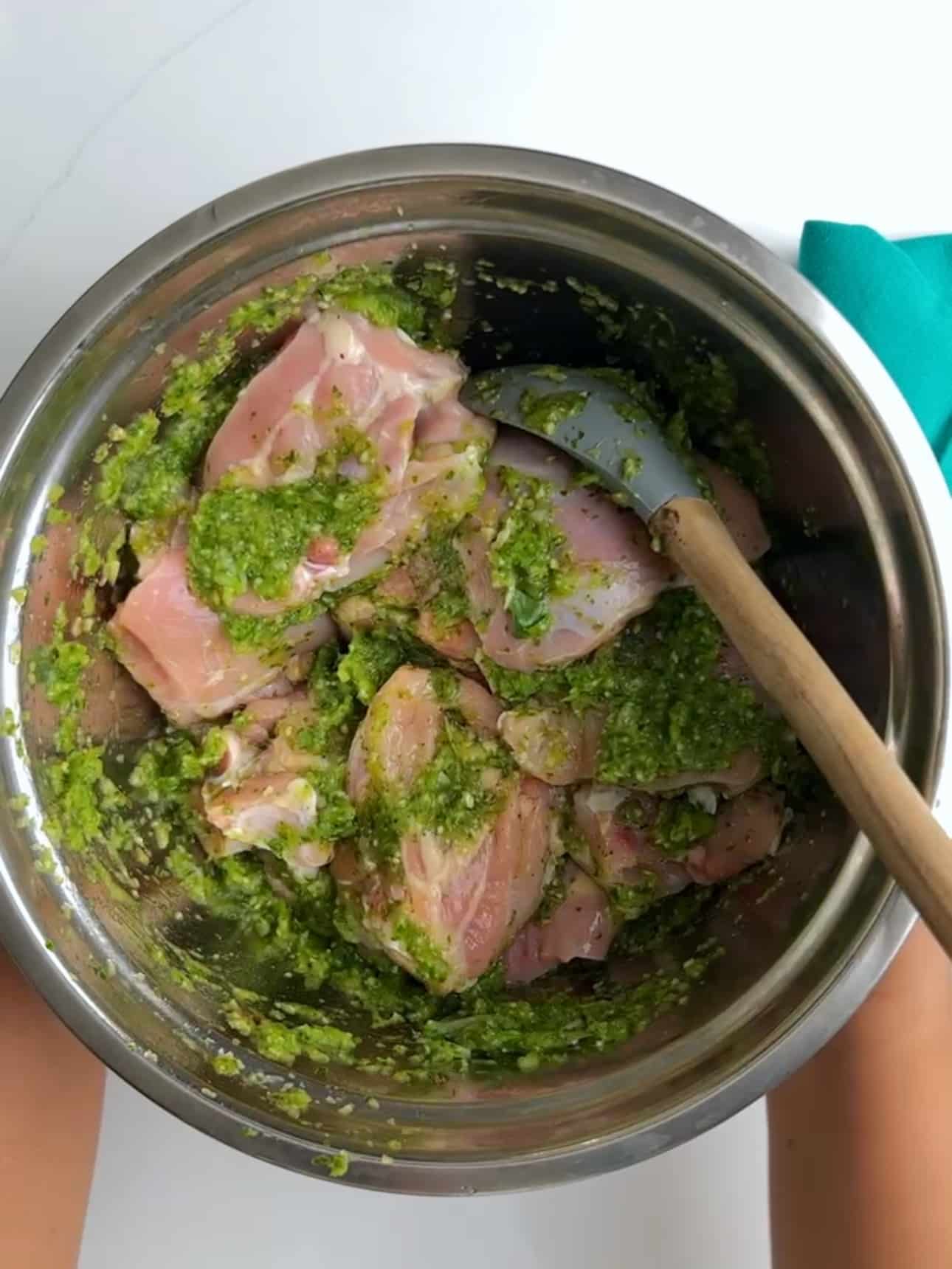 Raw chicken thighs coated in a green herb seasoning mixture in a metal mixing bowl.