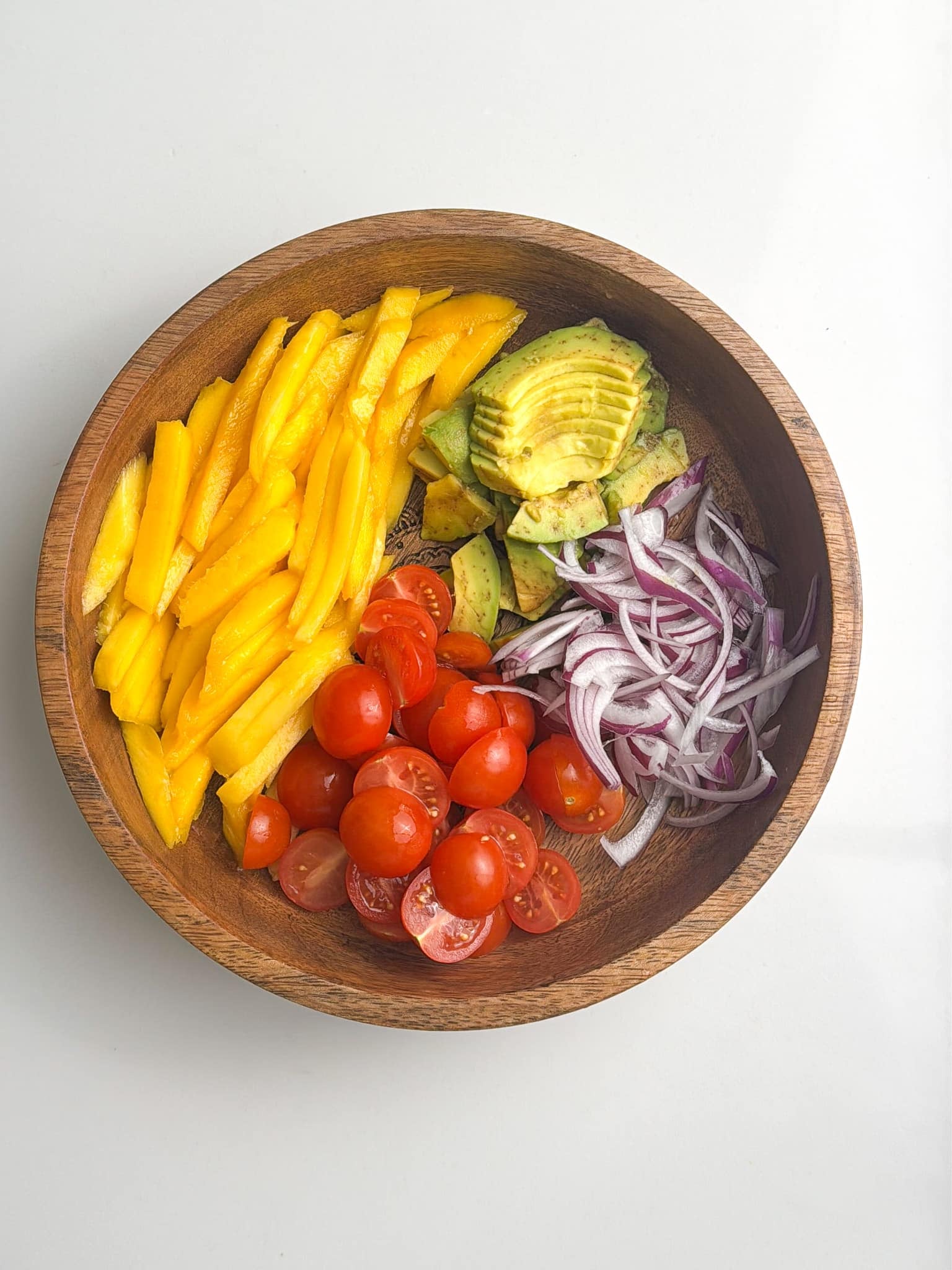 A wooden bowl arranged with sliced mango, avocado, cherry tomatoes, and red onion before mixing.