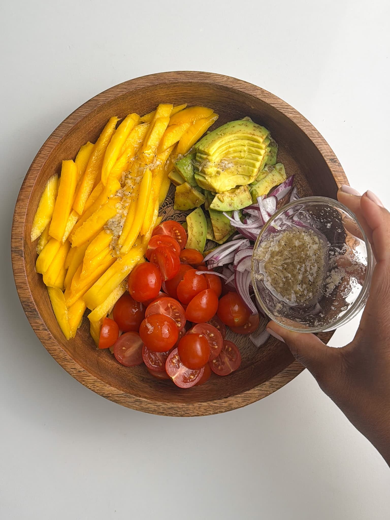 A hand pouring vinaigrette over a bowl of sliced mango, avocado, cherry tomatoes, and red onion.