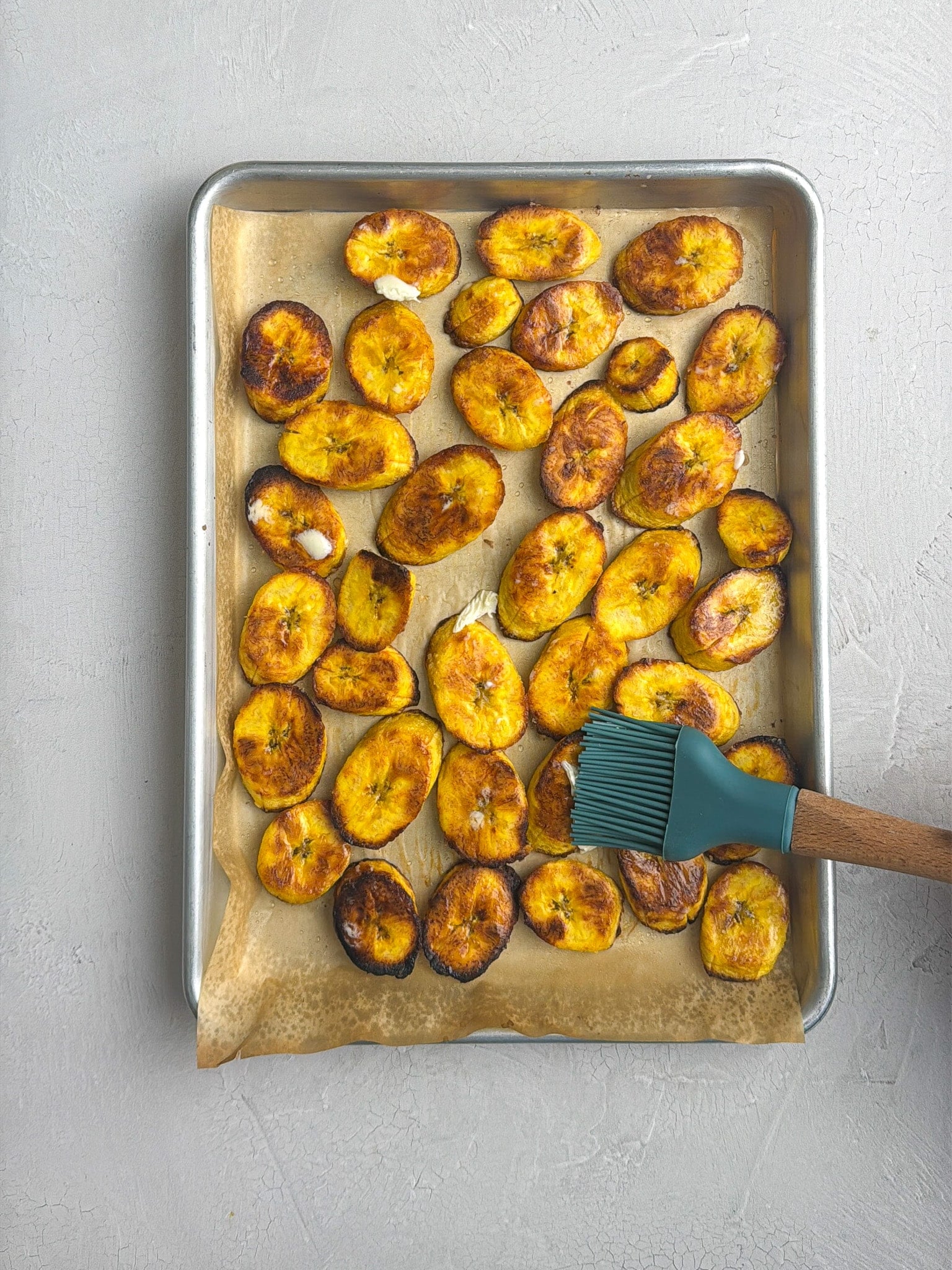 A hand brushes roasted plantain slices with butter on a baking sheet.