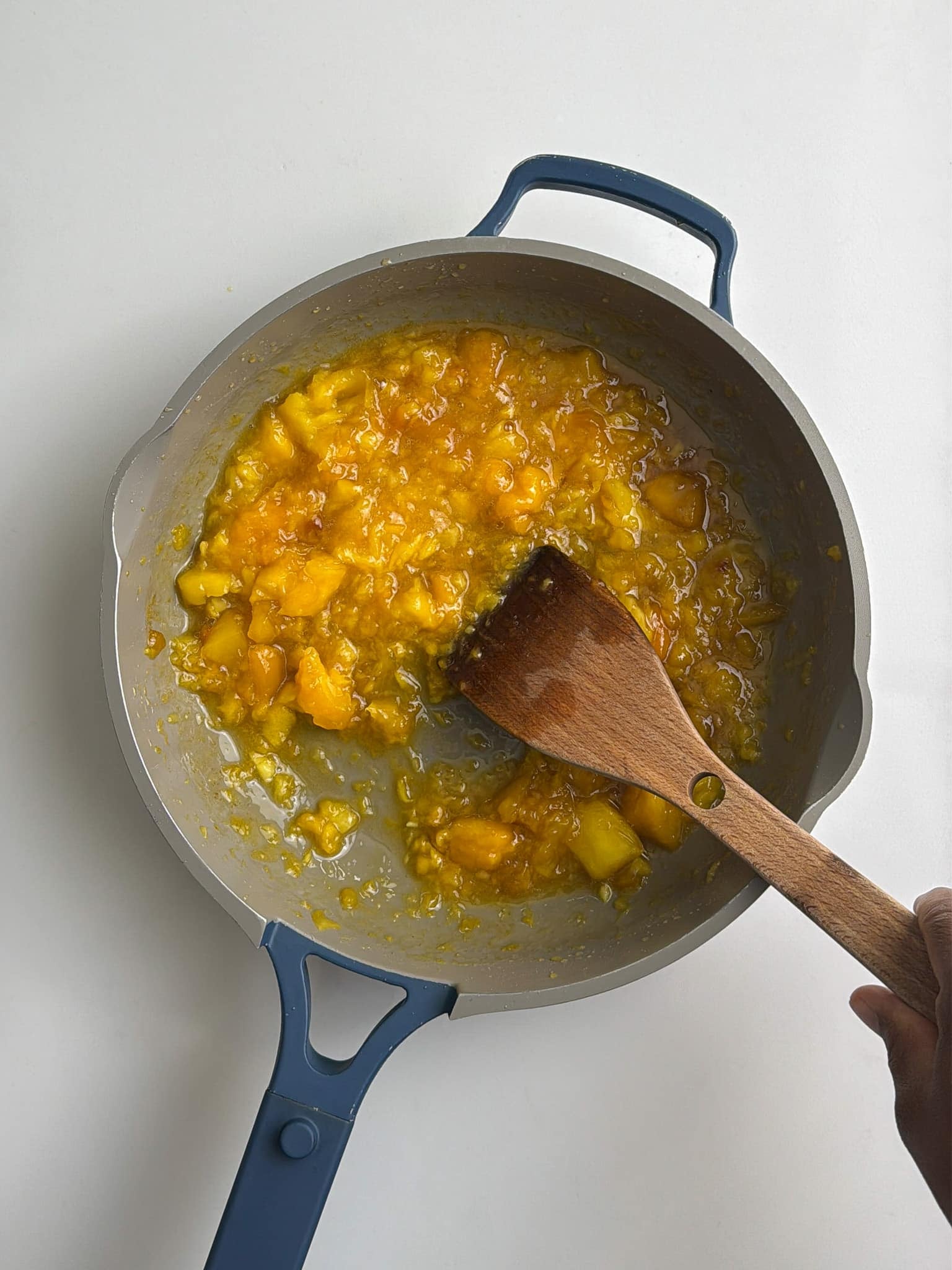 Diced mango cooking down in a saucepan while being stirred with a wooden spoon.