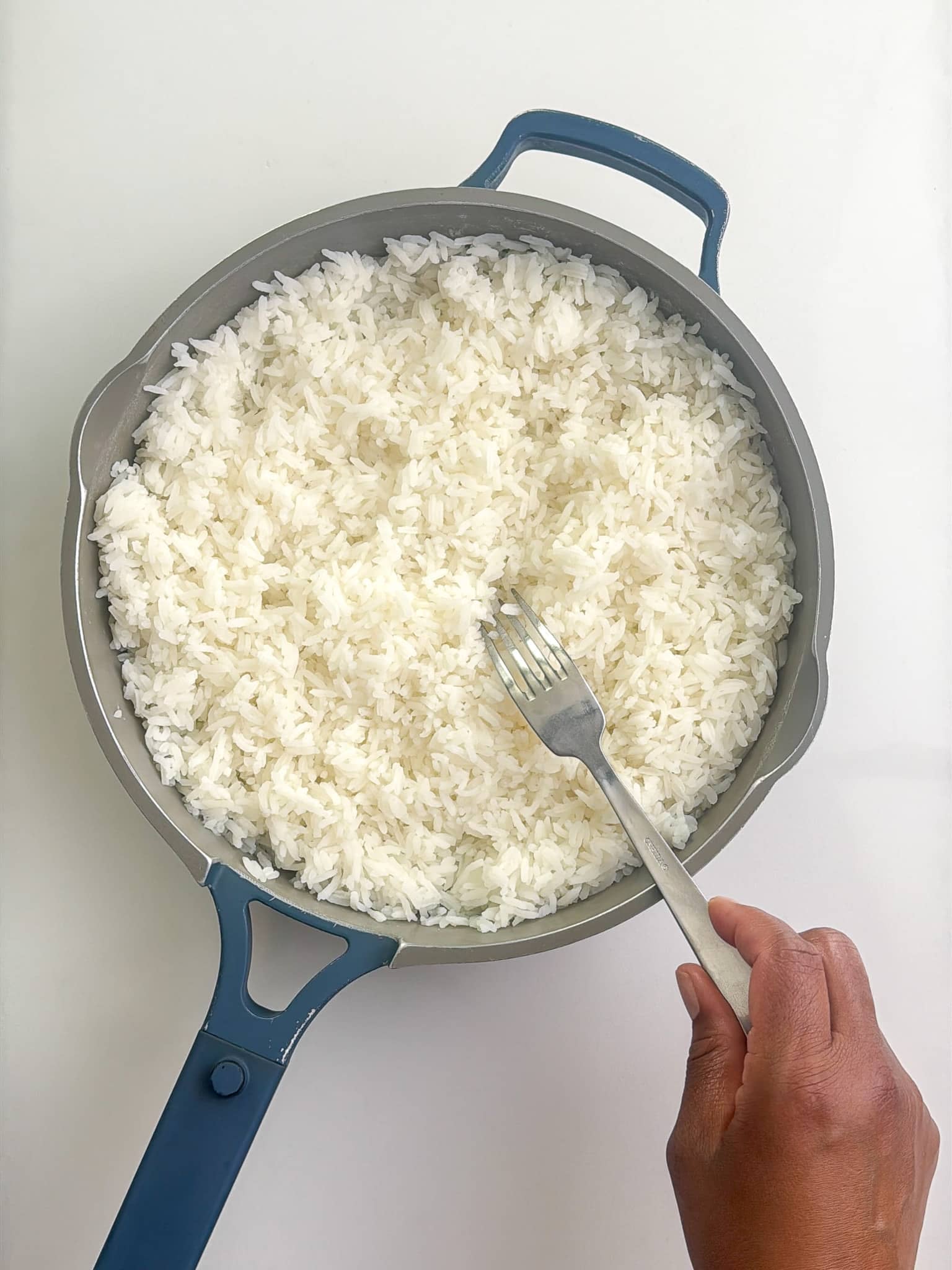 Hand using a fork to fluff freshly cooked white rice in a pot, separating the grains.