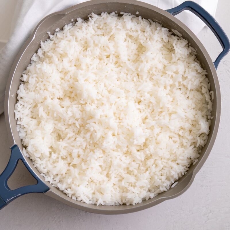A pot of freshly cooked white rice with fluffy, separate grains, shown in a wide pan on a light surface.