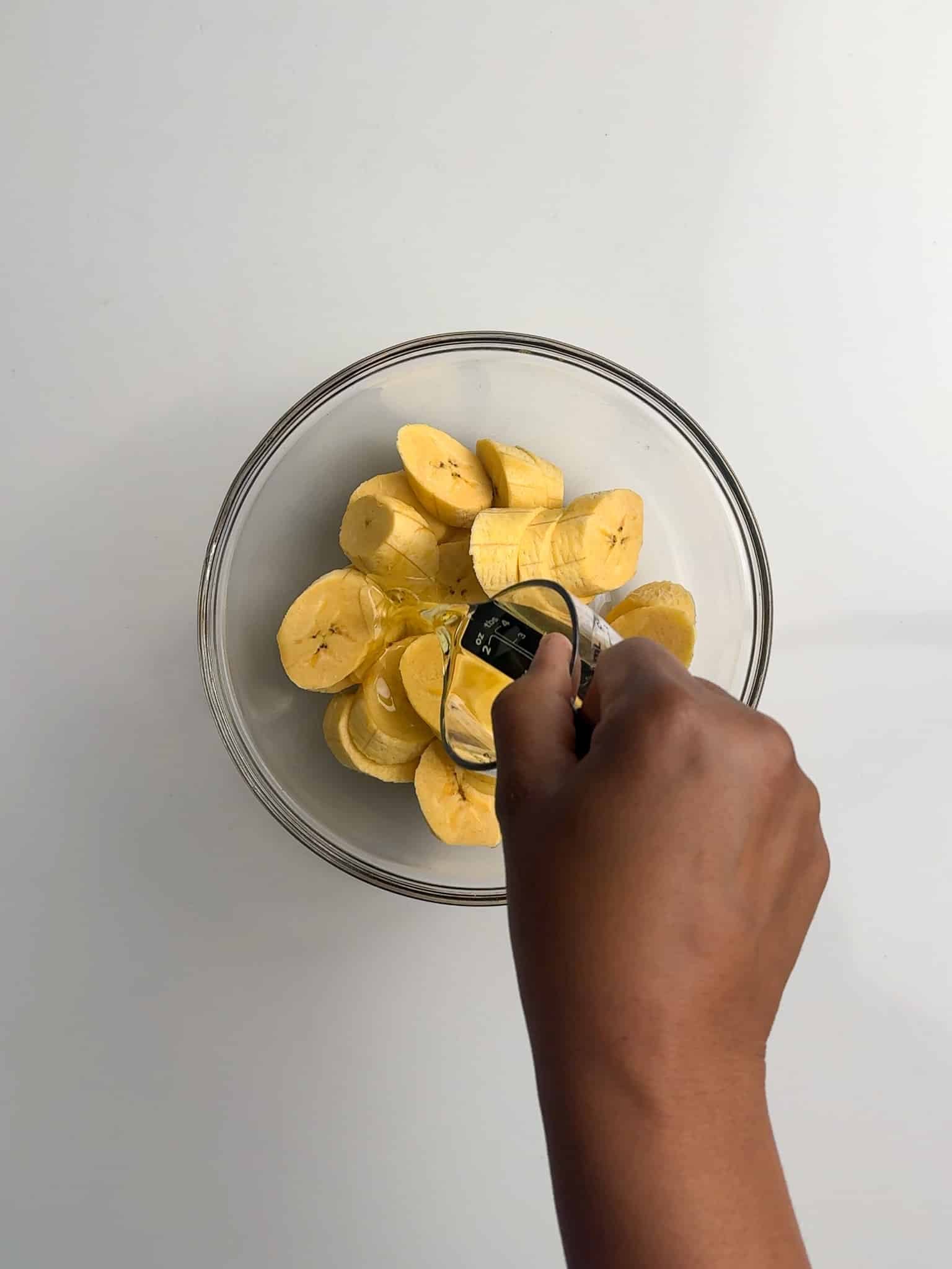 A hand pours oil over sliced plantains in a glass bowl.