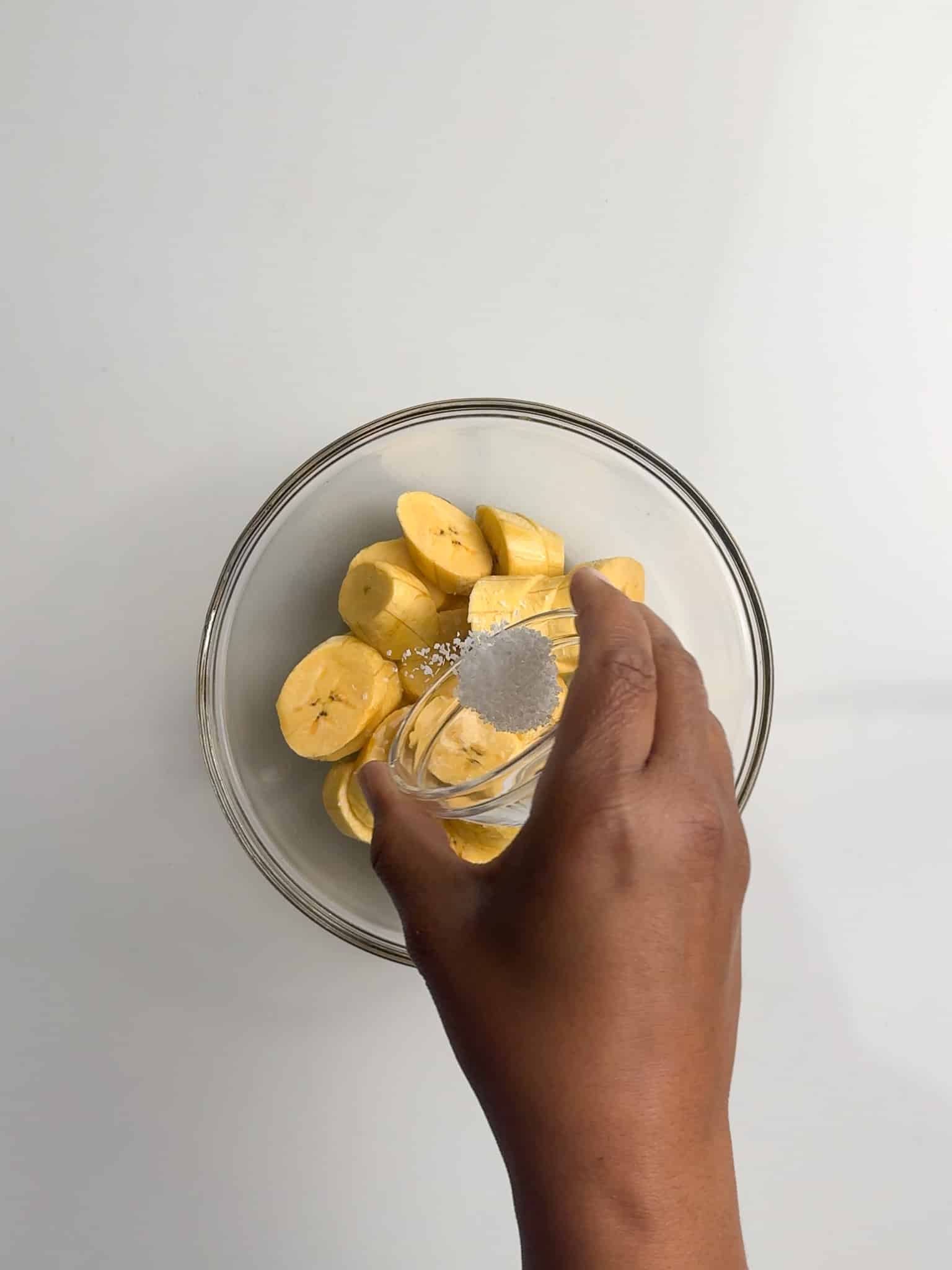 A hand sprinkles salt over sliced plantains in a glass bowl.