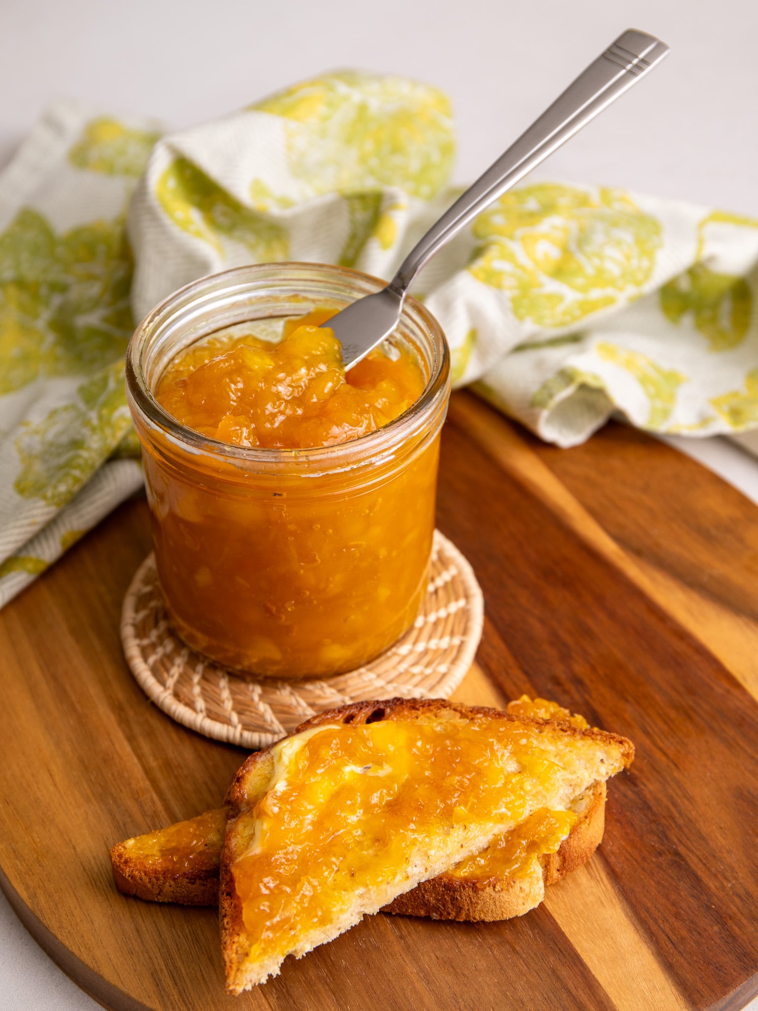 A spoon scooping mango jam from a jar with toast in the background.