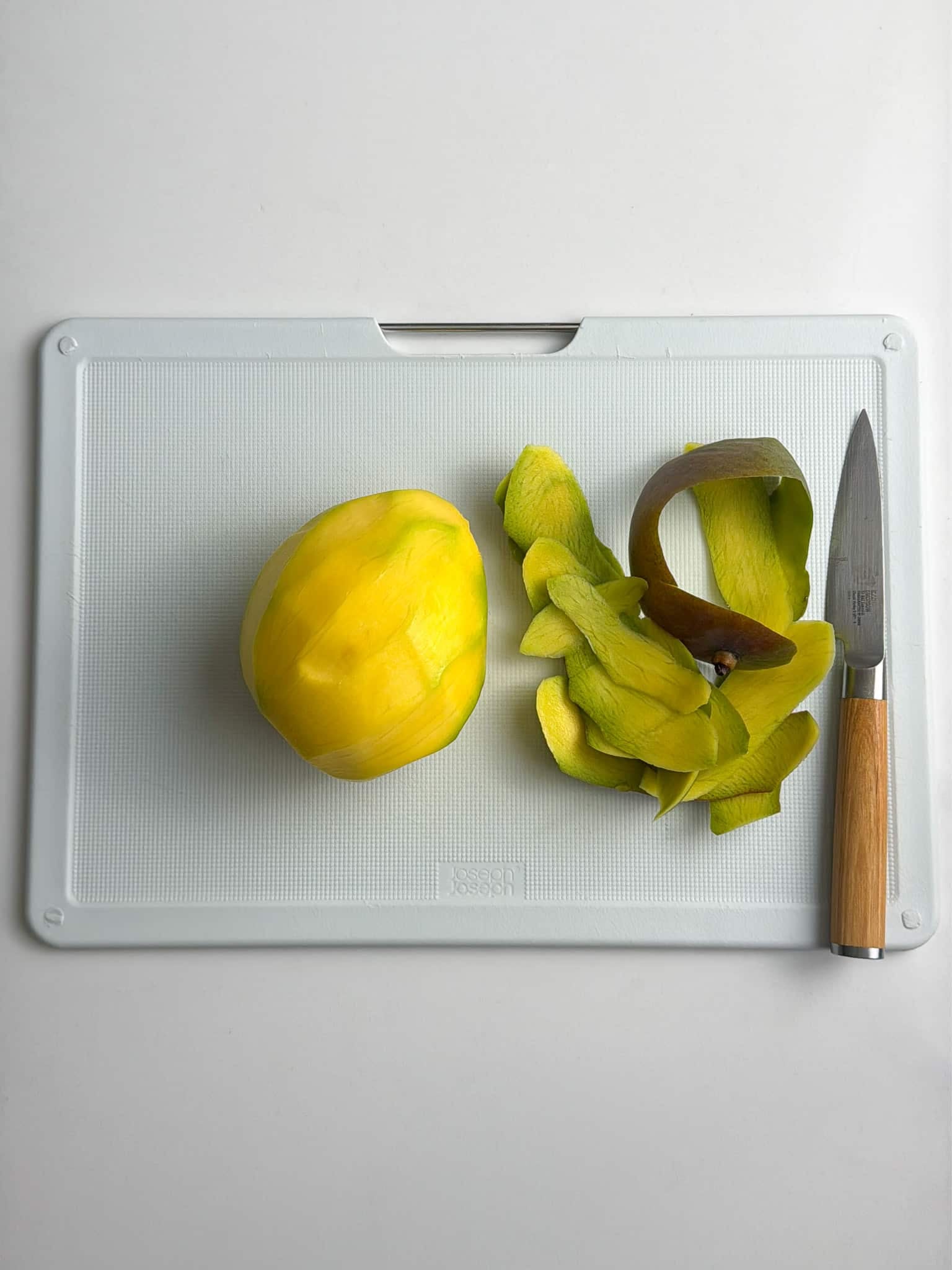 Peeled green mango with skin scraps and a knife on a cutting board.