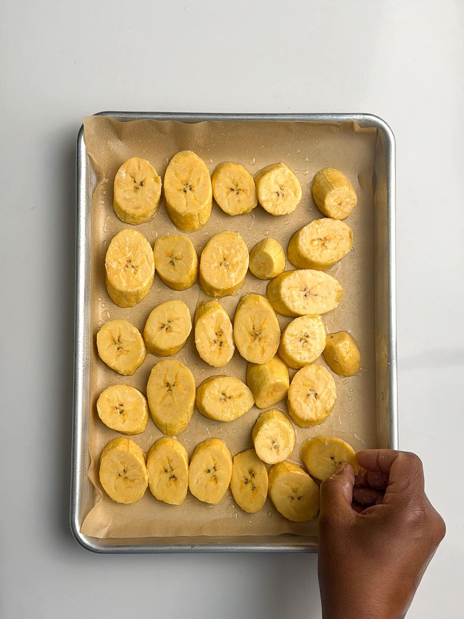 A hand places sliced plantains in a single layer on a parchment-lined baking sheet.
