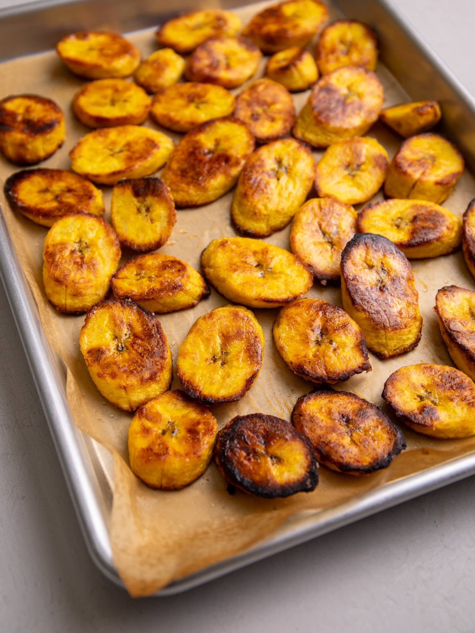 Baked plantain slices with caramelized edges are spread across a parchment-lined baking sheet.