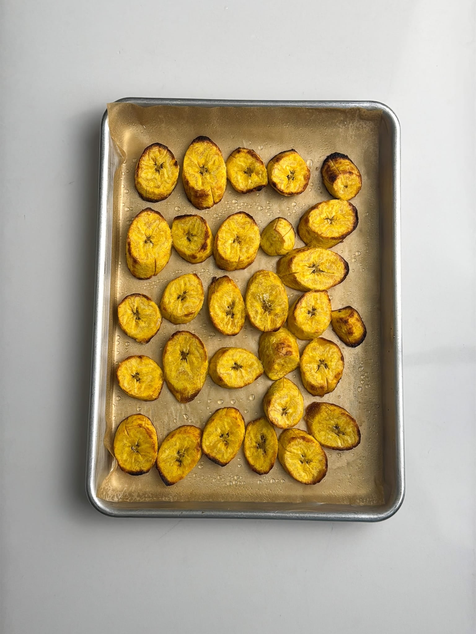 Roasted plantain slices spread across a baking sheet with golden brown edges.