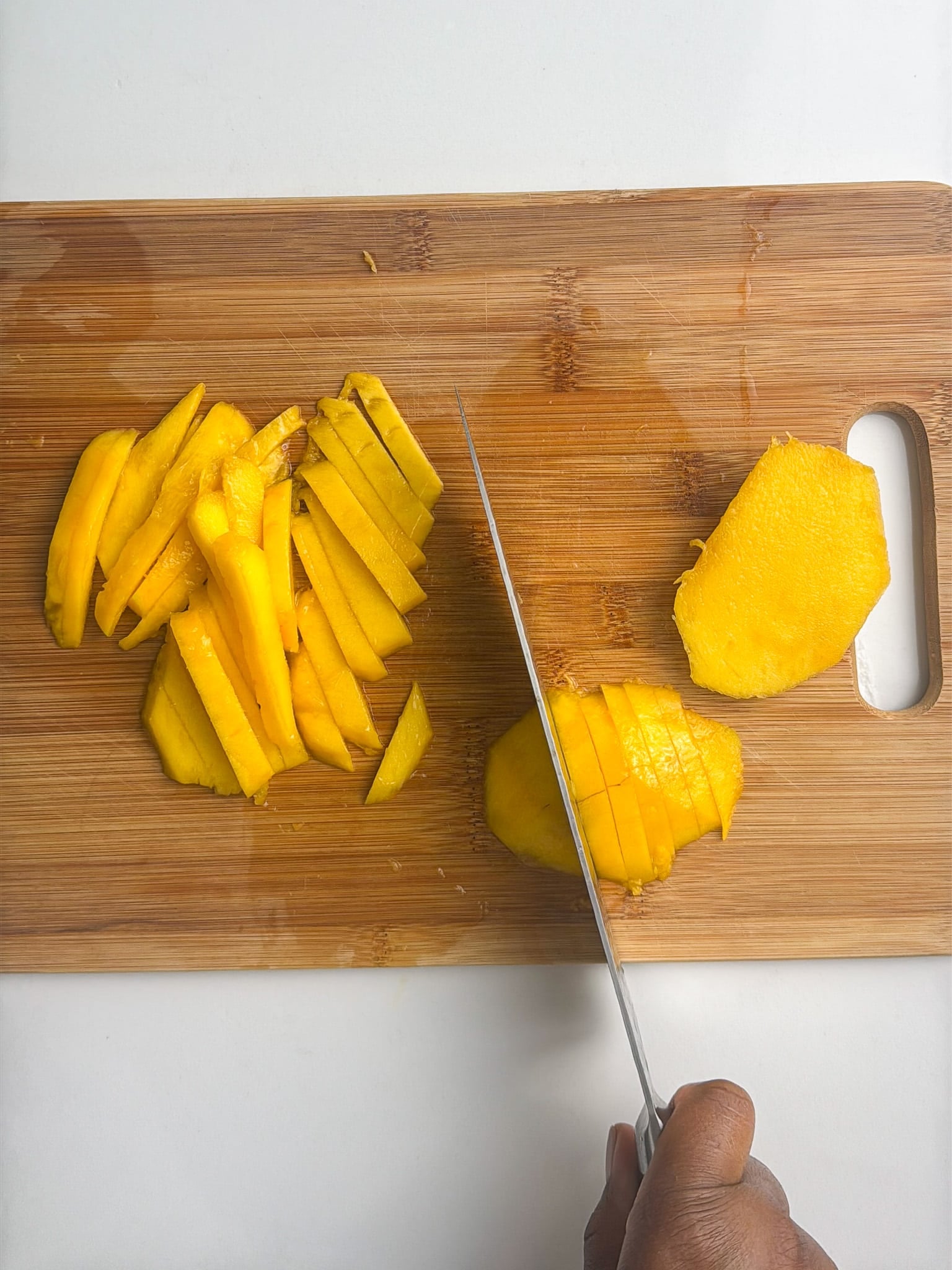 Fresh mango being sliced into thin strips on a wooden cutting board.