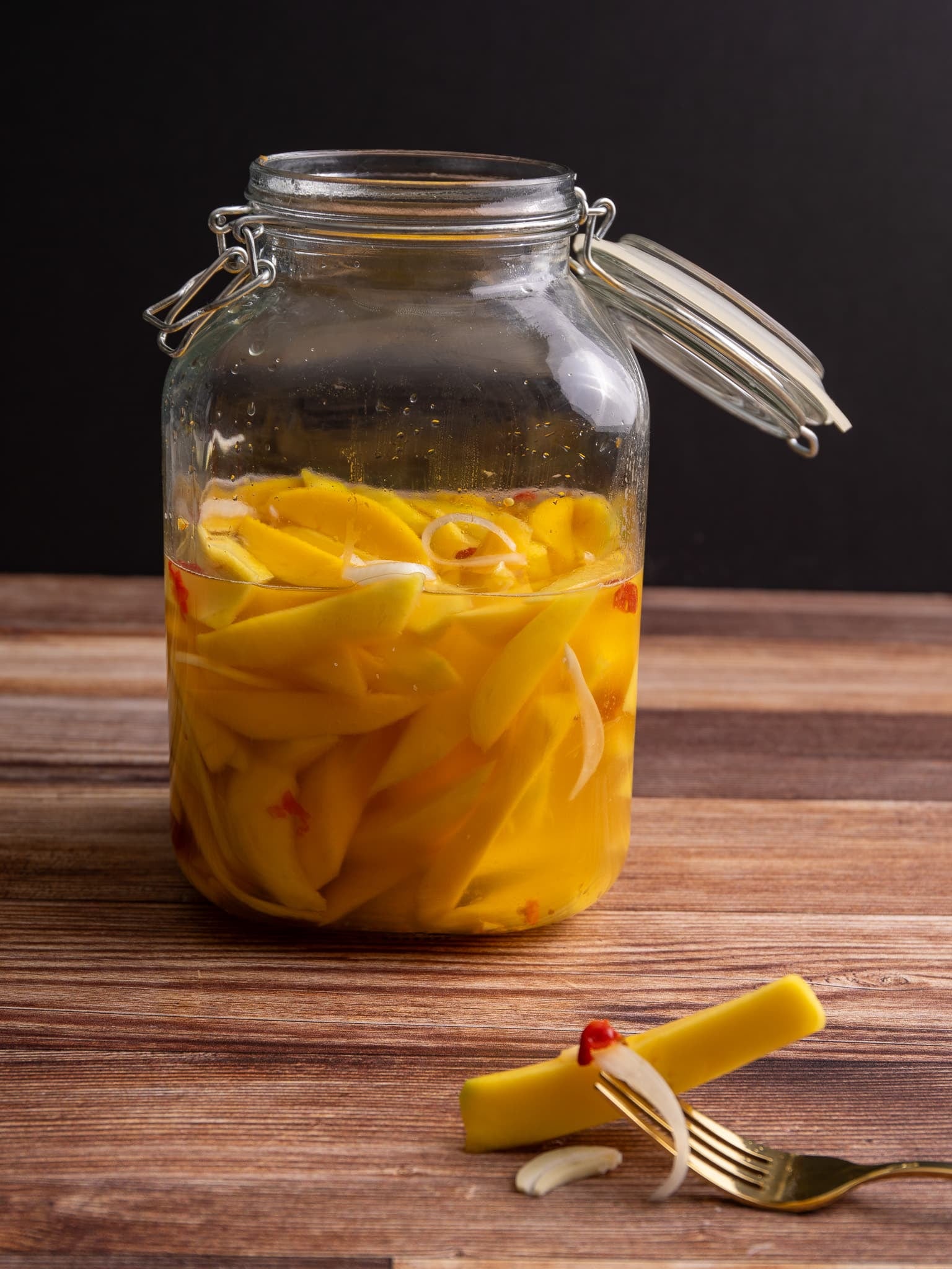 Glass jar of pickled mango on a wooden surface with brine and spices visible.