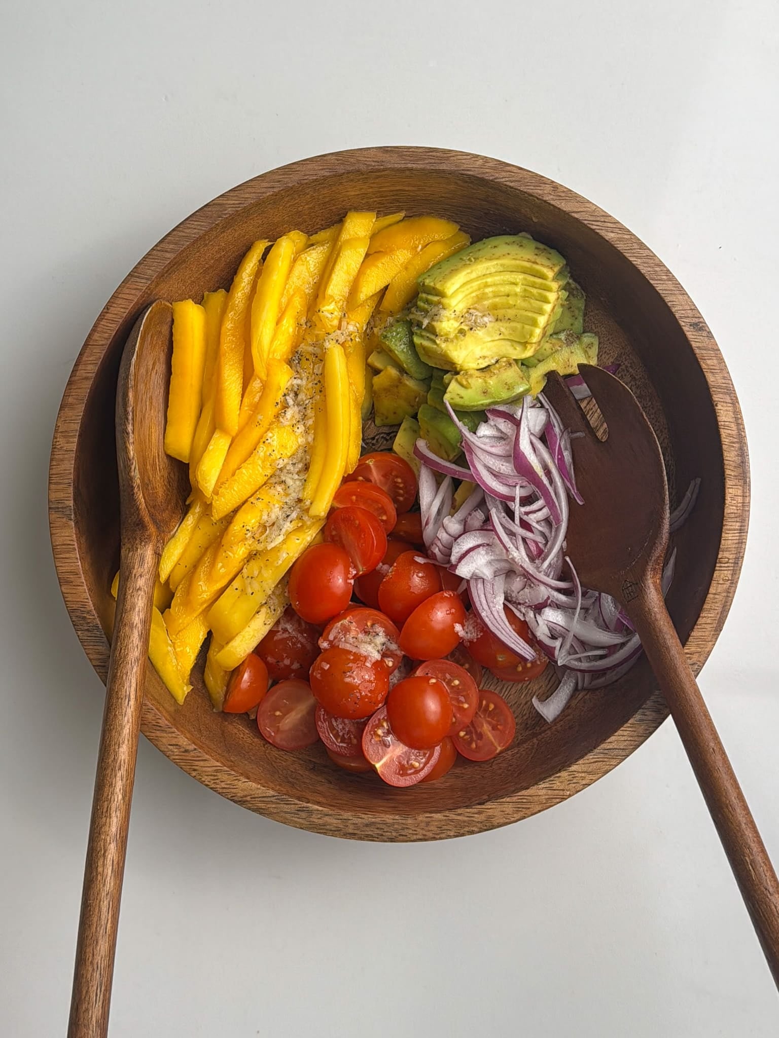 A wooden bowl filled with sliced mango, avocado, cherry tomatoes, and red onion being mixed with a wooden spoon.