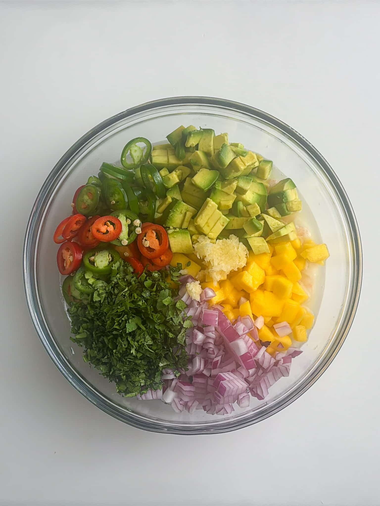 Fresh shrimp ceviche ingredients including avocado, mango, red onion, cilantro, jalapeños, and garlic in a glass bowl.