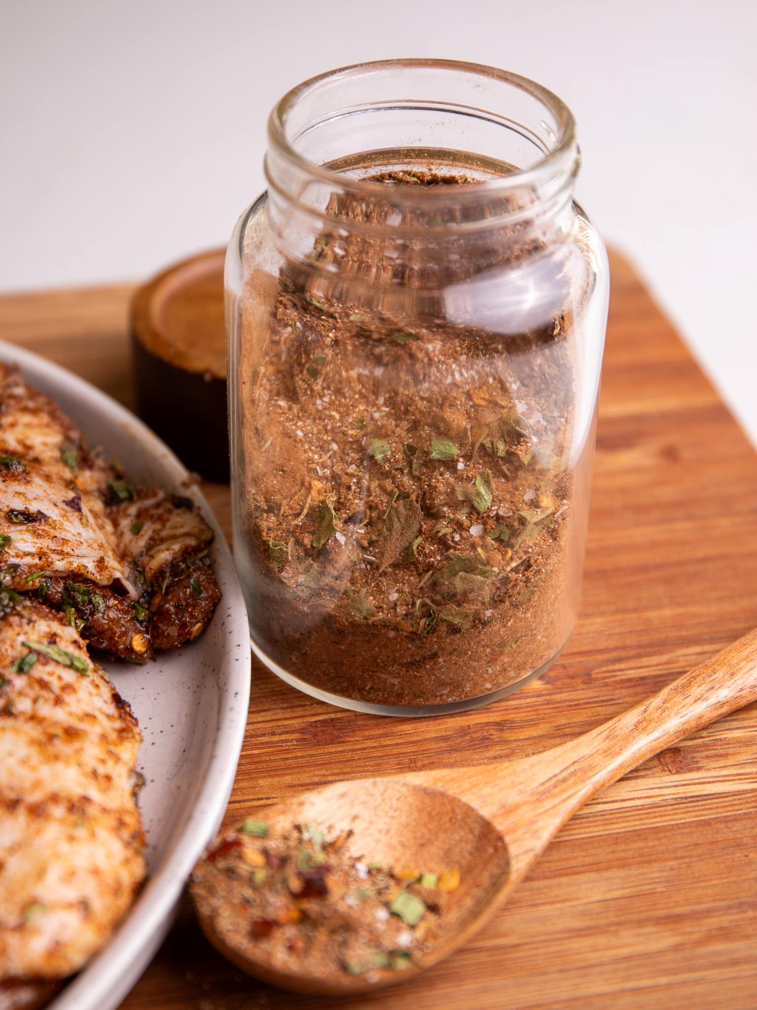 Glass jar filled with homemade Jamaican jerk seasoning dry rub beside cooked jerk chicken and a wooden spoon of seasoning.