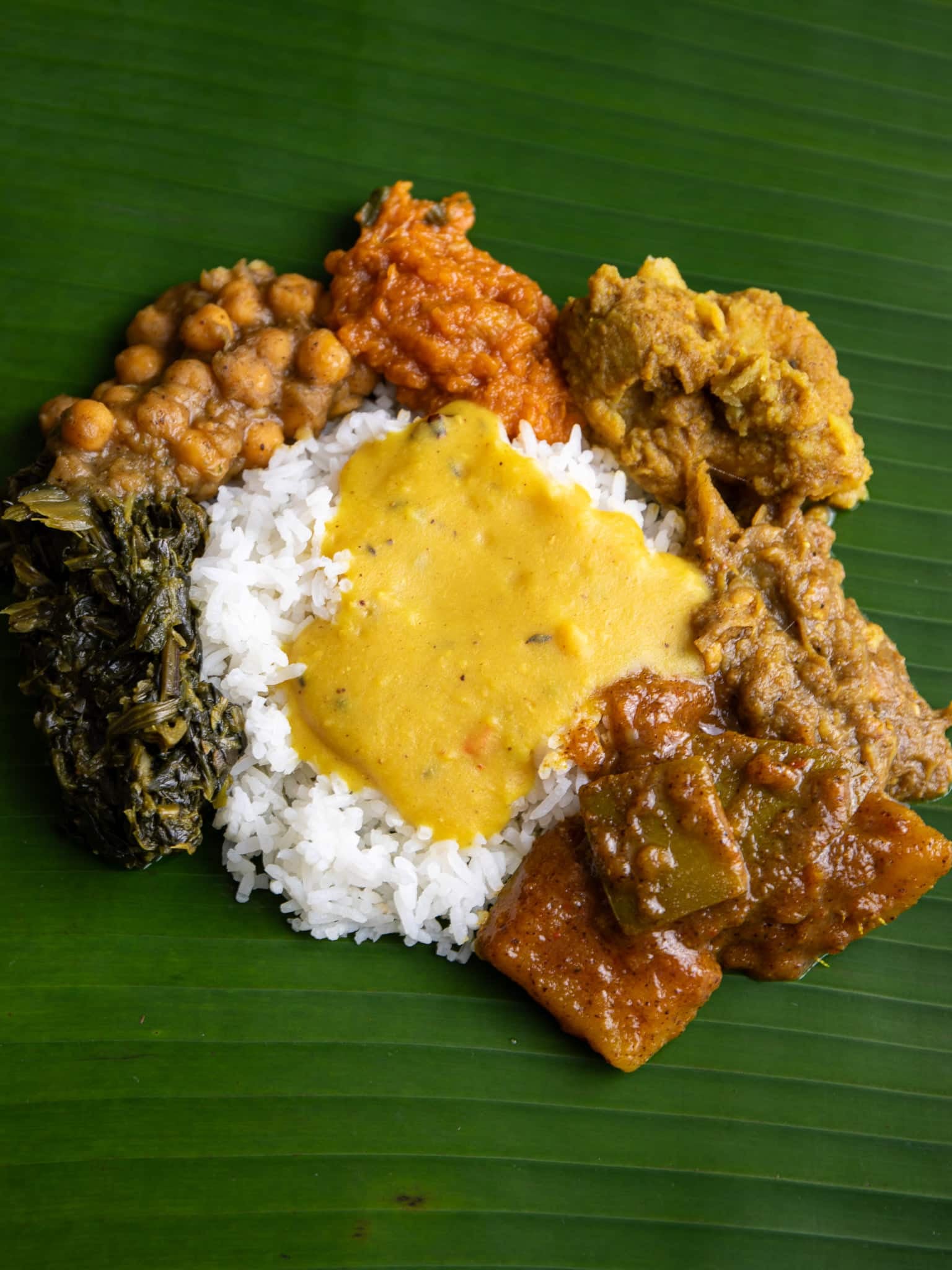 Guyanese seven curry on a banana leaf: dhal over white rice, surrounded by bhaji, channa, pumpkin, katahar, mango curry and more
